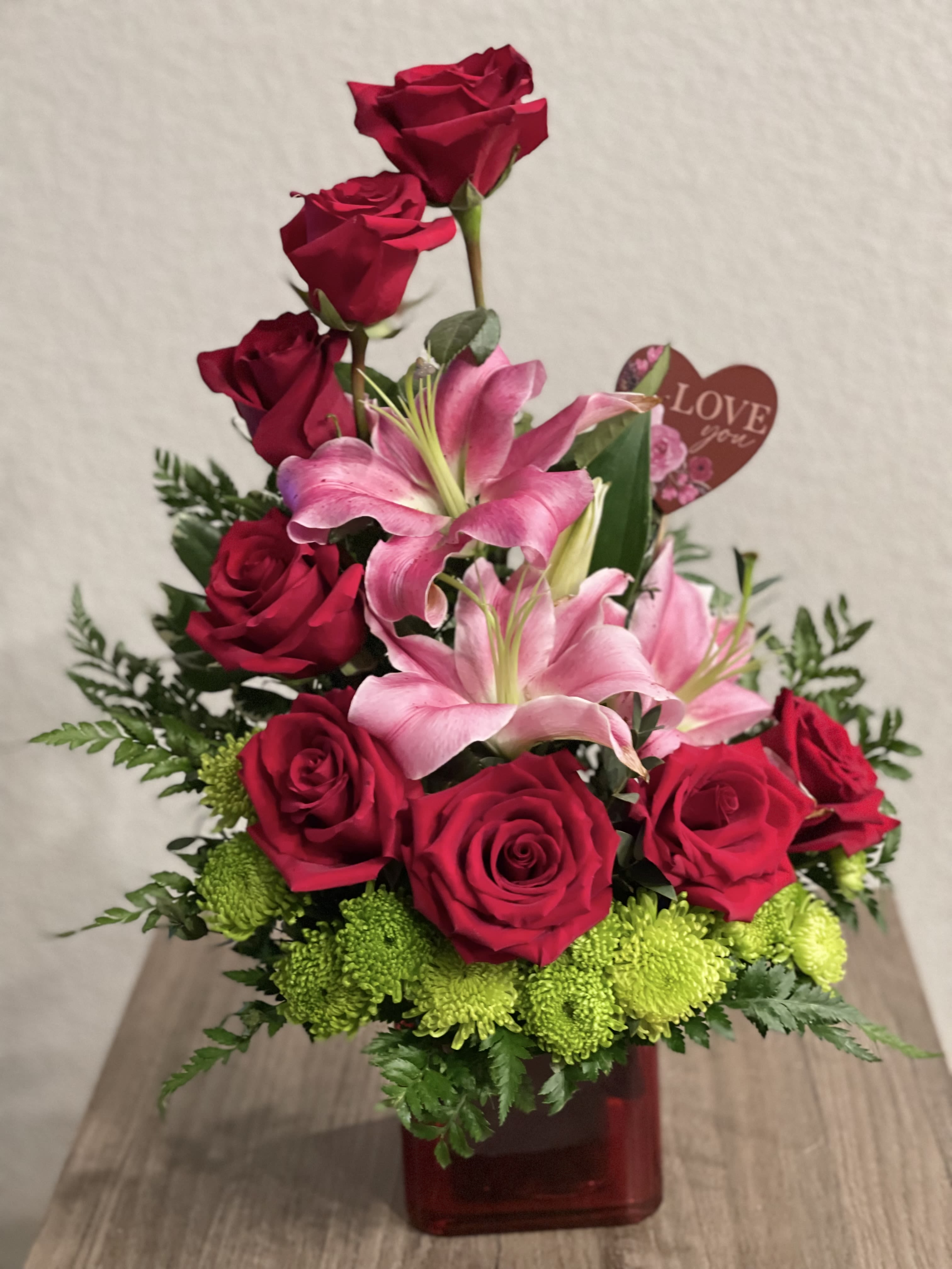 Red roses and pink lilies arranged in a red vase with a heart tag