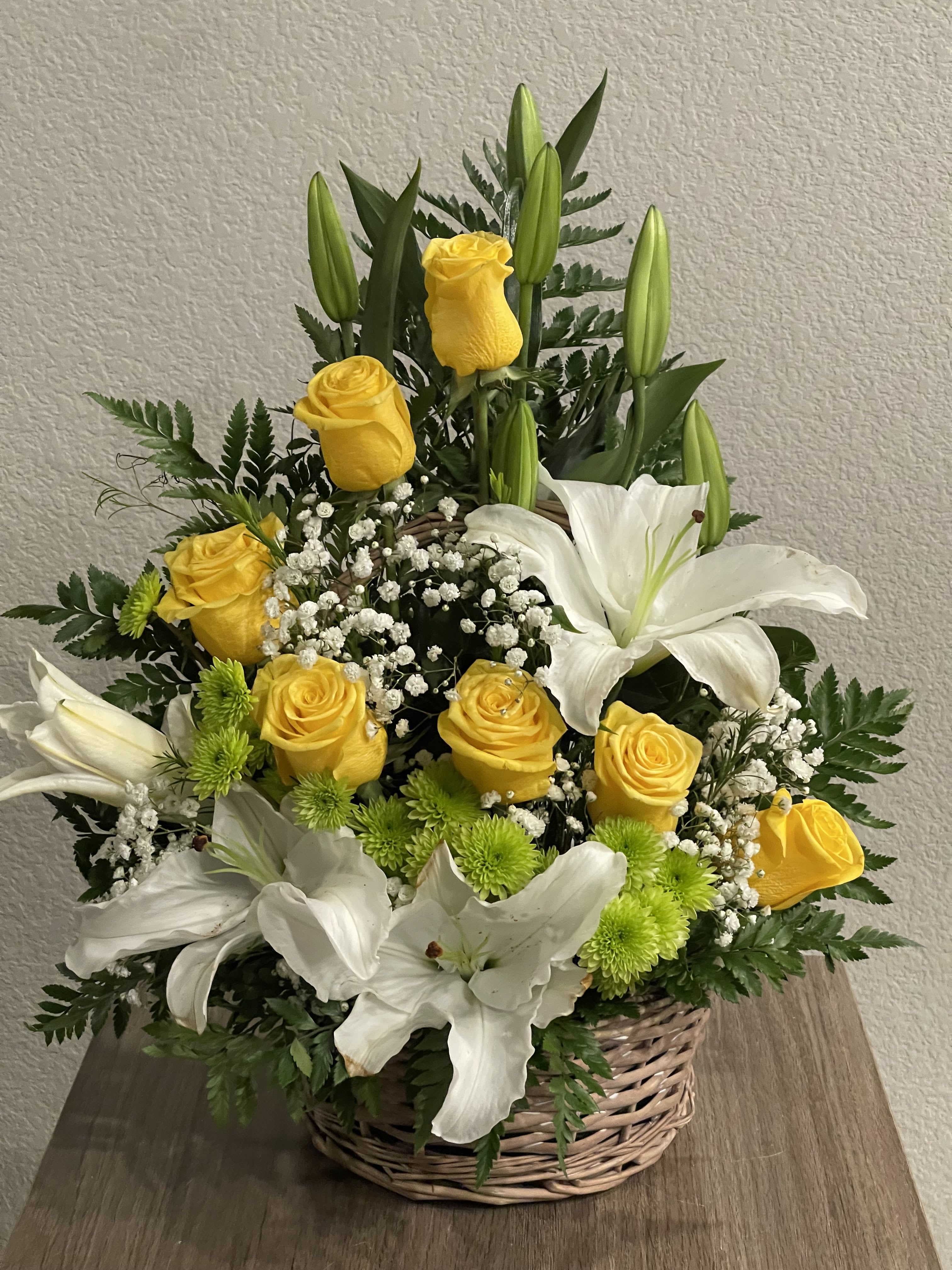 Home sweet home  - Sunlit yellow roses and large white lilies unite in a wicker basket for a bright, cheerful birthday bouquet or thoughtful thank-you gift. The "Home sweet home" arrangement reads fresh and uplifting-perfect for a spring celebration or to bring warmth to someone settling into a new place-while the white-and-yellow contrast keeps the feel elegantly composed.  This full, layered design features upright lily buds rising above rounded rose blooms, clusters of small white filler flowers, and vivid lime-green button chrysanthemums that pop near the base. Dark green foliage adds depth and structure, and the woven basket gives a rustic, homey finish that's ready to display.  Choose this for someone who should feel welcomed, appreciated, or gently celebrated-whether it's a congratulations, a birthday, or a get-well moment. The basket arrives arranged and ready to place on a table or entryway; lilies and roses benefit from fresh water and a cool spot to extend bloom life.