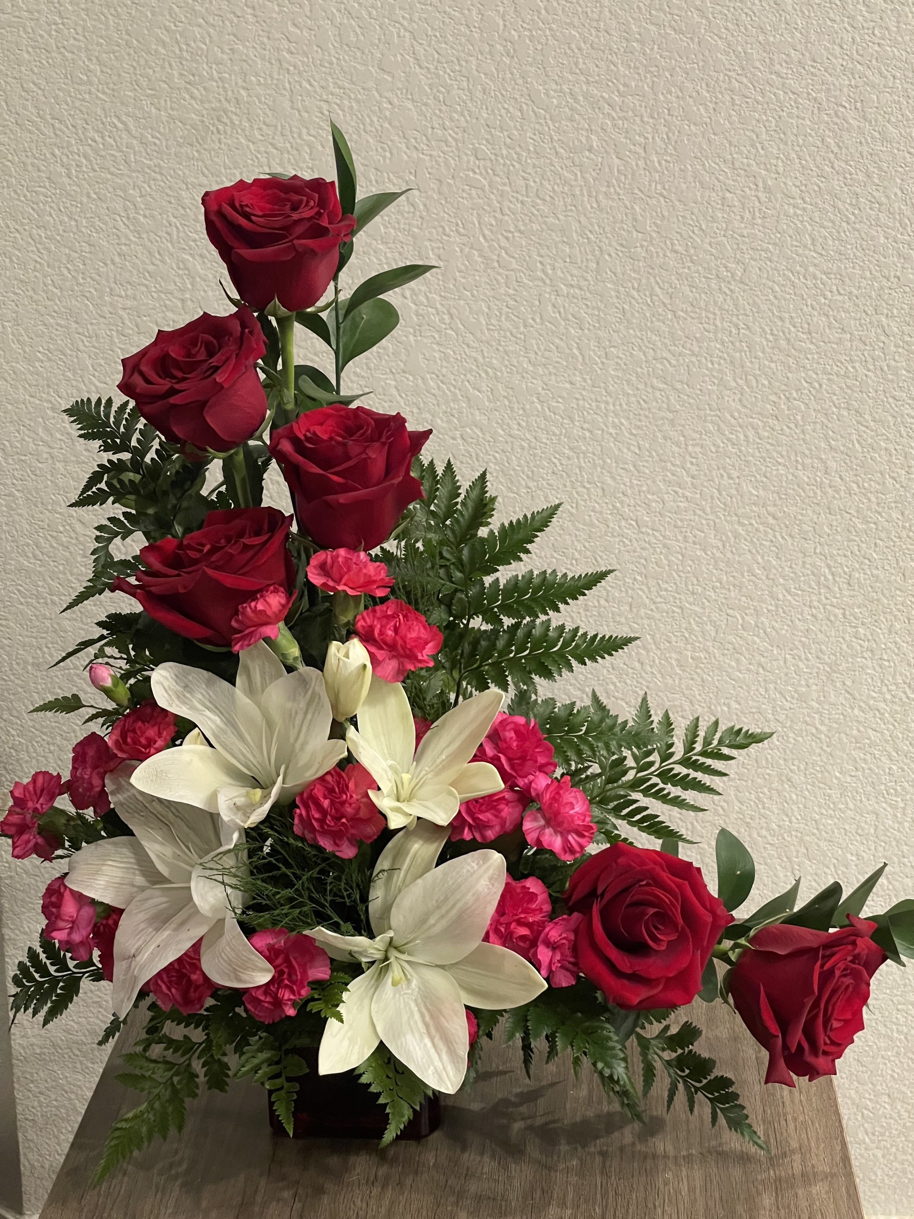 Grace  Bloom  - Tall red roses lift the eye above clusters of large white lilies and bright pink carnations in this romantic anniversary bouquet - a red, white, pink and green arrangement designed for celebrations of love or moments of heartfelt sympathy. The vertical lines of the roses give graceful movement while the white lilies provide striking contrast; the pink carnations add a tender, soft touch so the recipient feels both admired and comforted.  Flowers are densely packed with fern-like greenery for a full, structured silhouette and are presented in a low rectangular vase that keeps the overall design compact and refined. Several rose stems extend slightly higher for height, while the lilies and carnations form rich layers at mid and low levels, making this an elegant choice for anniversaries, Valentine's Day, or as a thoughtful sympathy tribute. Care: keep water fresh, recut stems occasionally, and place away from direct heat to prolong bloom life.