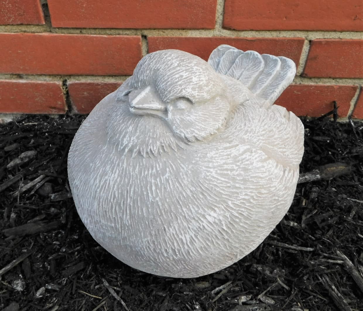 White bird-shaped garden statue resting on dark mulch in front of a brick wall