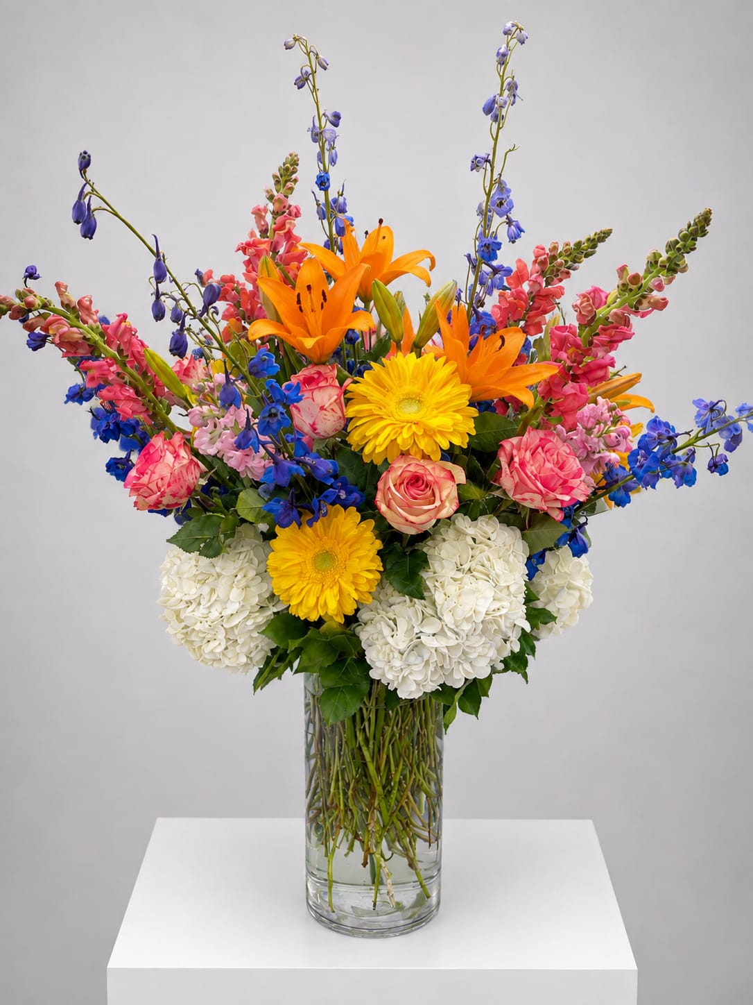 Tall mixed bouquet in a clear glass vase with orange lilies, yellow daisies, pink roses, and blue flowers