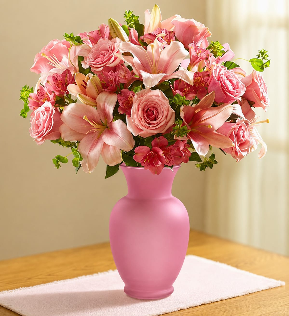 Sunlit Pink Elegance - A lush cluster of blush and rose tones makes this bouquet feel like sunlight captured in petals - perfect as an anniversary bouquet or a romantic gift. This flower arrangement for birthdays, Mother's Day, or simply to say "I'm thinking of you" centers on pink roses, pale pink lilies, and deeper pink alstroemeria arranged in a rounded, abundant shape that reads soft, feminine, and cheerful.  The composition is gathered in a matte pink vase that continues the monochromatic palette and gives the display a polished, decorative finish. Layered blooms create gentle texture and visual depth: velvety roses, trumpet-shaped lilies, and delicate, freckled alstroemeria mingle with greenery for contrast. The arrangement's size and fullness make it ideal for spring and summer celebrations, new baby visits, cheering someone getting well, or offering comfort at a sympathy or funeral service.  This is the kind of floral gift that will make the recipient feel truly seen-celebrated on a birthday, soothed during recovery, or honored on Women's Day or Breast Cancer Awareness Month. Care is simple: keep water fresh and place in indirect light to extend bloom life; choose your size at checkout to match the occasion and the room you want to brighten.
