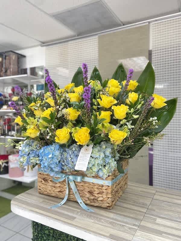 Large basket of yellow roses, blue hydrangeas, purple flowers and bold green leaves with a blue ribbon.