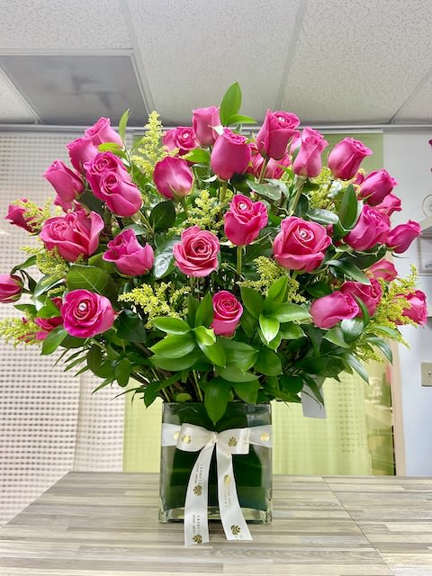 Tall arrangement of bright pink roses with filler flowers in a square glass vase with a white ribbon