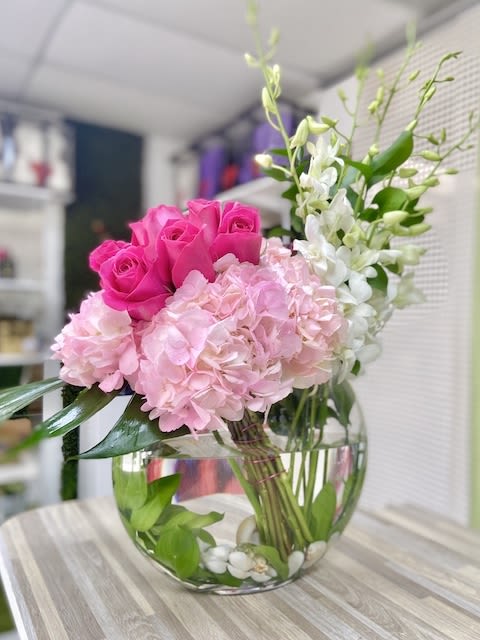 Round glass vase with pink roses, pink hydrangeas, and white orchids arranged low with visible stems and stones.