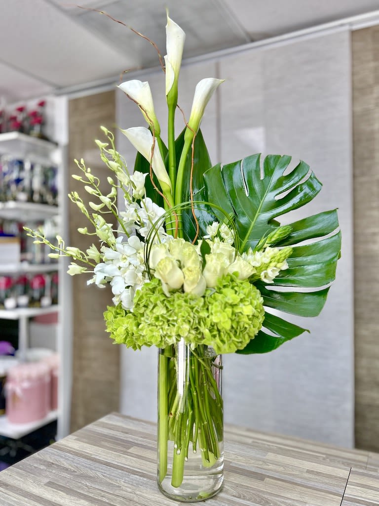 Tall white and green arrangement with calla lilies, orchids, and hydrangeas in a clear glass cylinder vase