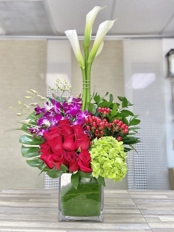 Tall modern arrangement with white calla lilies, red roses, purple orchids, and green hydrangea in a glass cube vase.