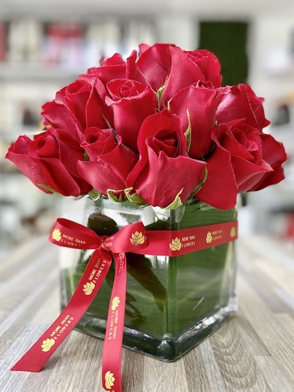 Compact arrangement of red roses in a clear glass cube vase with a red printed ribbon