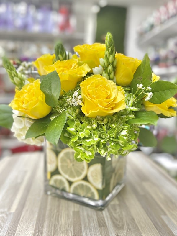 Square glass vase filled with lemon slices holding yellow roses and green hydrangeas with white accents.