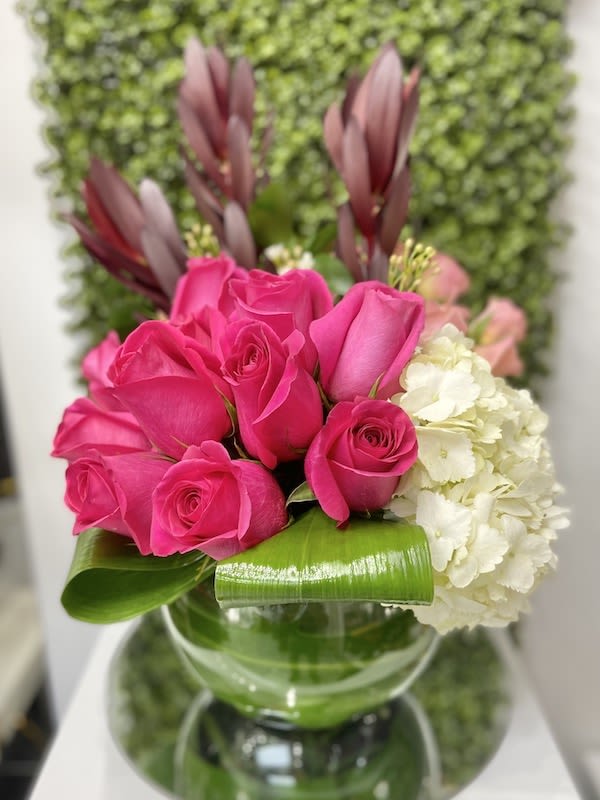 Low glass vase with hot pink roses, white hydrangea, and looped green leaves in a compact arrangement