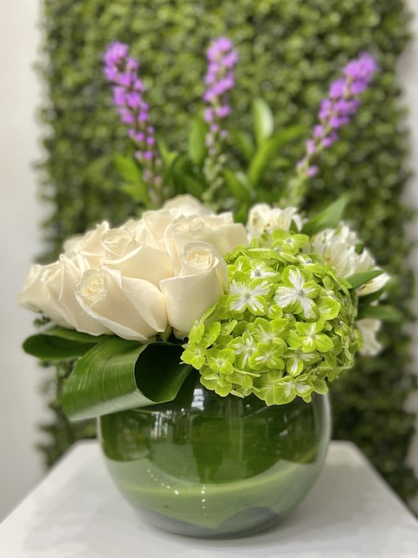 Low arrangement of white roses and green hydrangea in a round green glass vase with tall purple flower spikes.