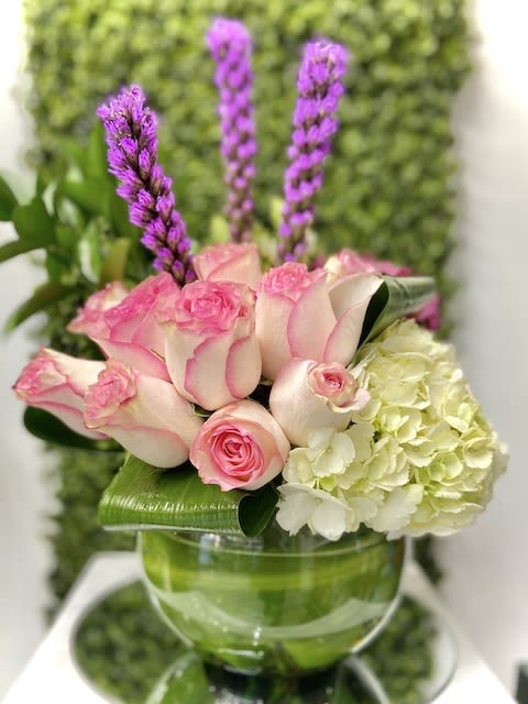 Low arrangement of pink roses, white hydrangea and tall purple spikes in a round glass bowl vase
