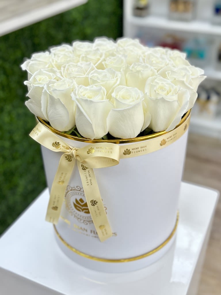 Round white hatbox filled with white roses and wrapped with a gold logo ribbon.
