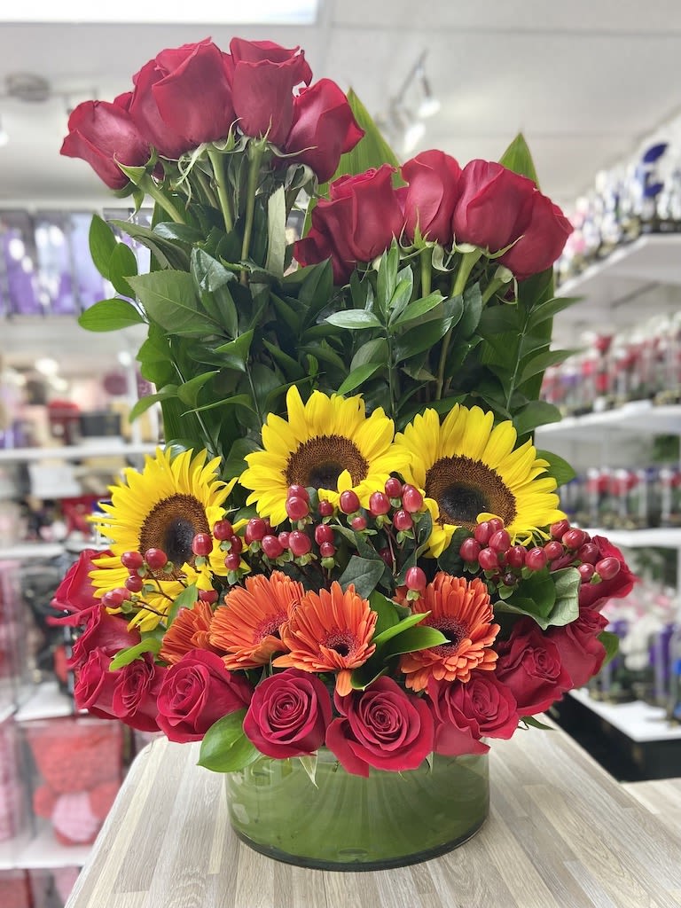 Tall arrangement of red roses, yellow sunflowers, and orange gerbera daisies in a round glass vase.