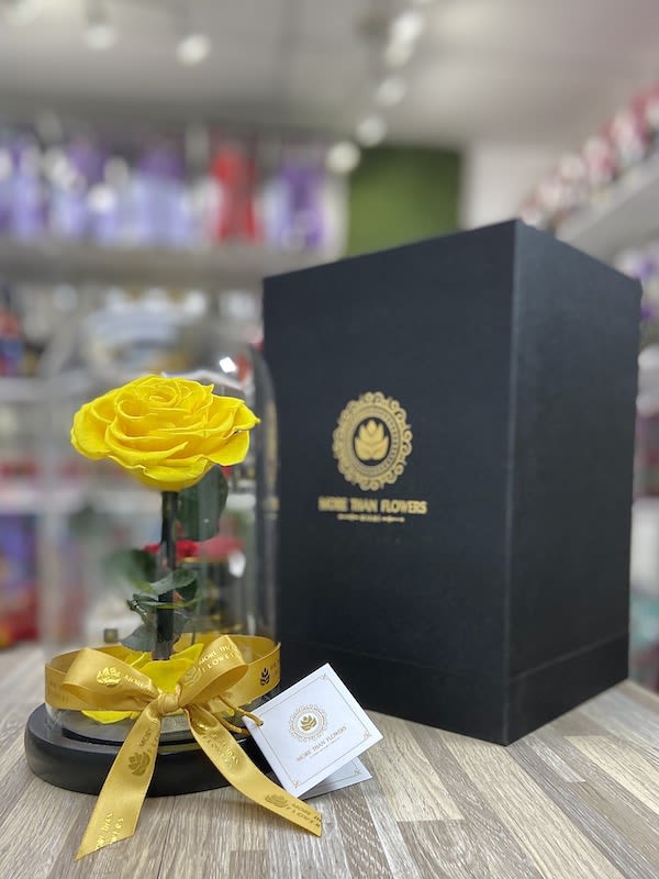 Single yellow rose under a glass dome with gold ribbon on black base beside a tall black gift box