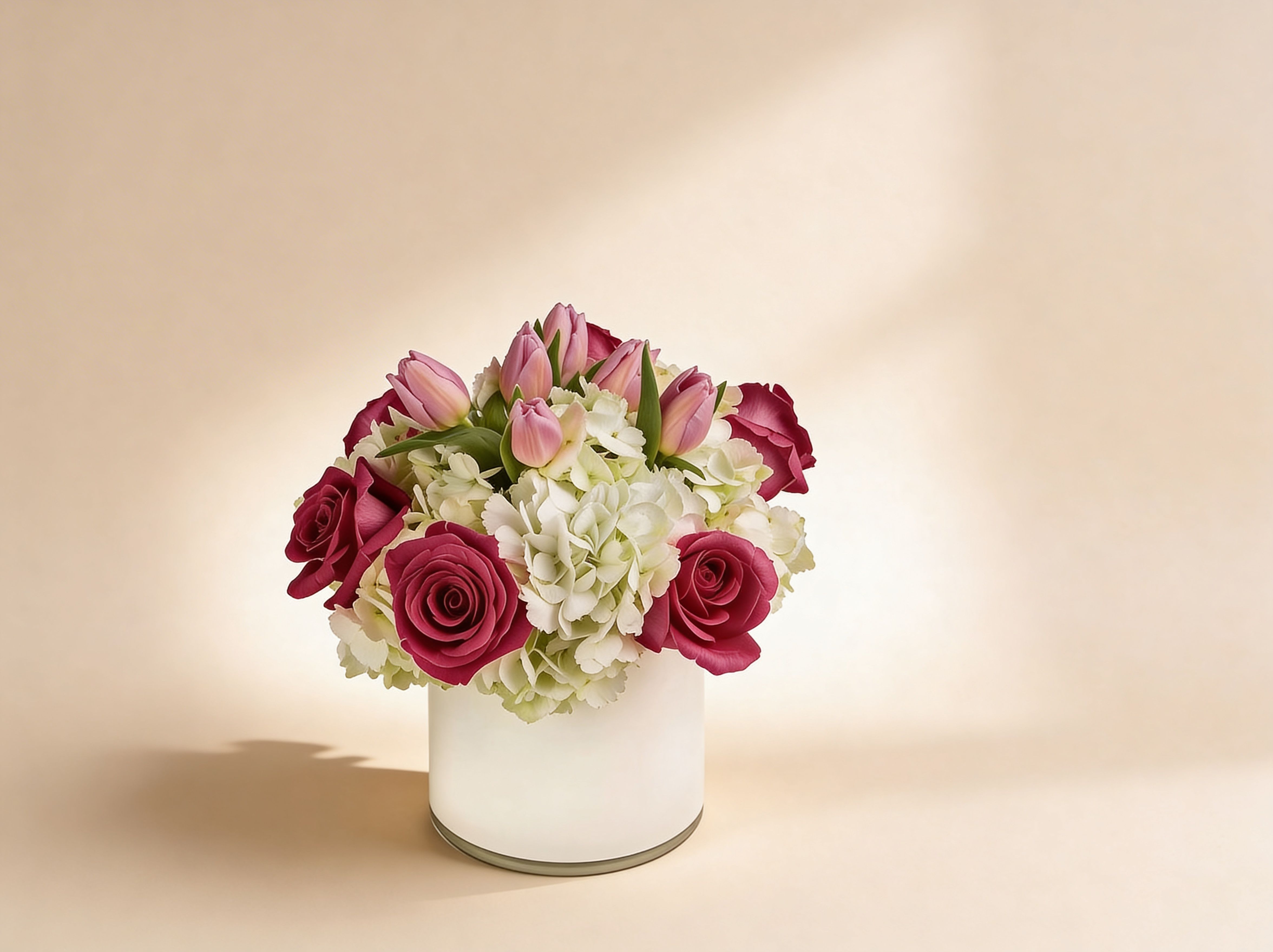 Low arrangement of pink roses, pink tulips, and white hydrangeas in a white cylinder vase