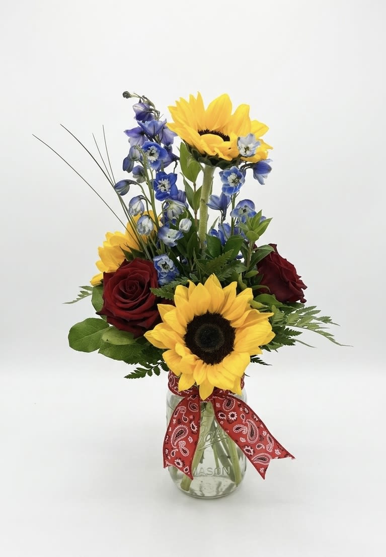 Hill Country Harmony - A Texas Classic for Any Celebration "Bring the soulful beauty of the Texas Hill Country right to their doorstep. Hill Country Harmony is a vibrant, textured arrangement that balances the bold, sunny energy of premium sunflowers with the deep, romantic velvet of red roses. Touches of midnight blue delphinium add a striking contrast, all gathered in a timeless mason jar and hand-finished with our signature red paisley bandana.  Whether you’re celebrating a milestone birthday, sending a heartfelt 'thank you,' or simply brightening a friend's day, this arrangement captures a spirit of warmth and authentic charm that fits every occasion perfectly."