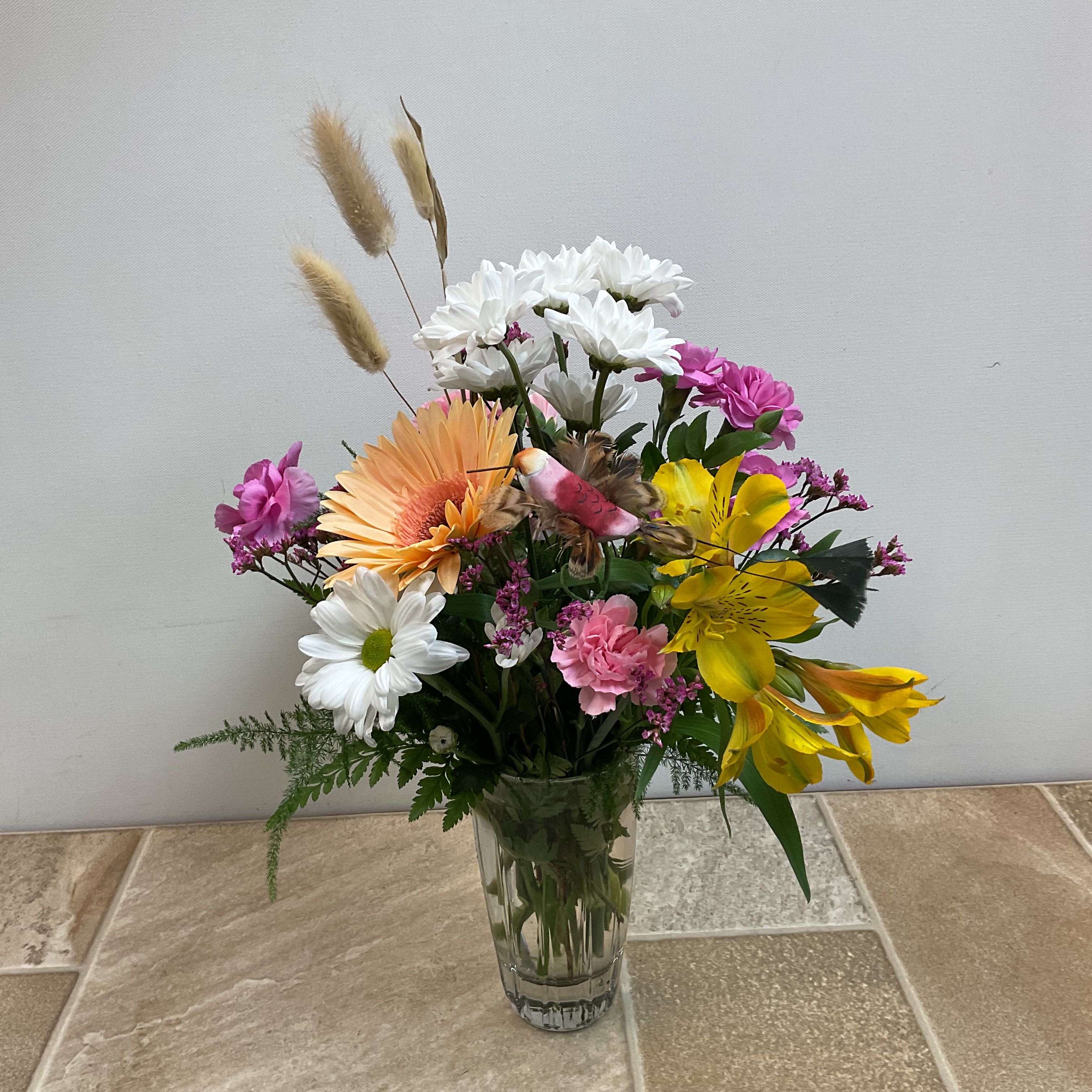 Hummingbird - Did you notice the pretty hummingbird figuring checking out the fresh flowers in this bouquet? This is a vibrant and cheerful floral arrangement presented in a glass vase. The bouquet features a mix of colors and textures, giving it a fresh and lively appearance. A large gerbera daisy takes center stage, surrounded by alstroemeria, mini carnations, and daisies. Accents include pink diamond limon and feathery green foliage.  Approximate size: 11” wide x 14” tall.   Primary flowers: daisy, lily, carnation