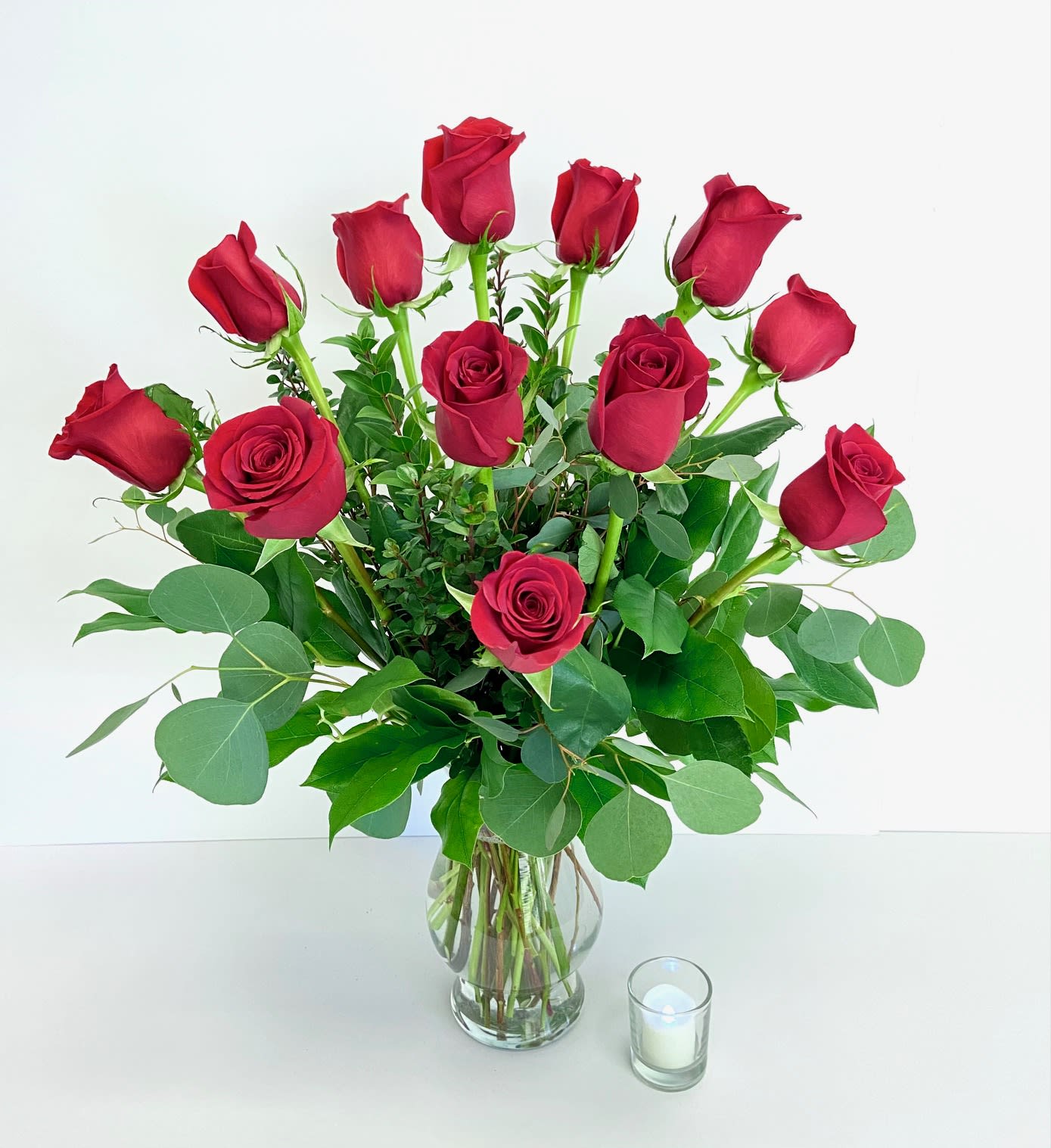 Red roses arranged in a clear glass vase with greenery and a small candle beside it