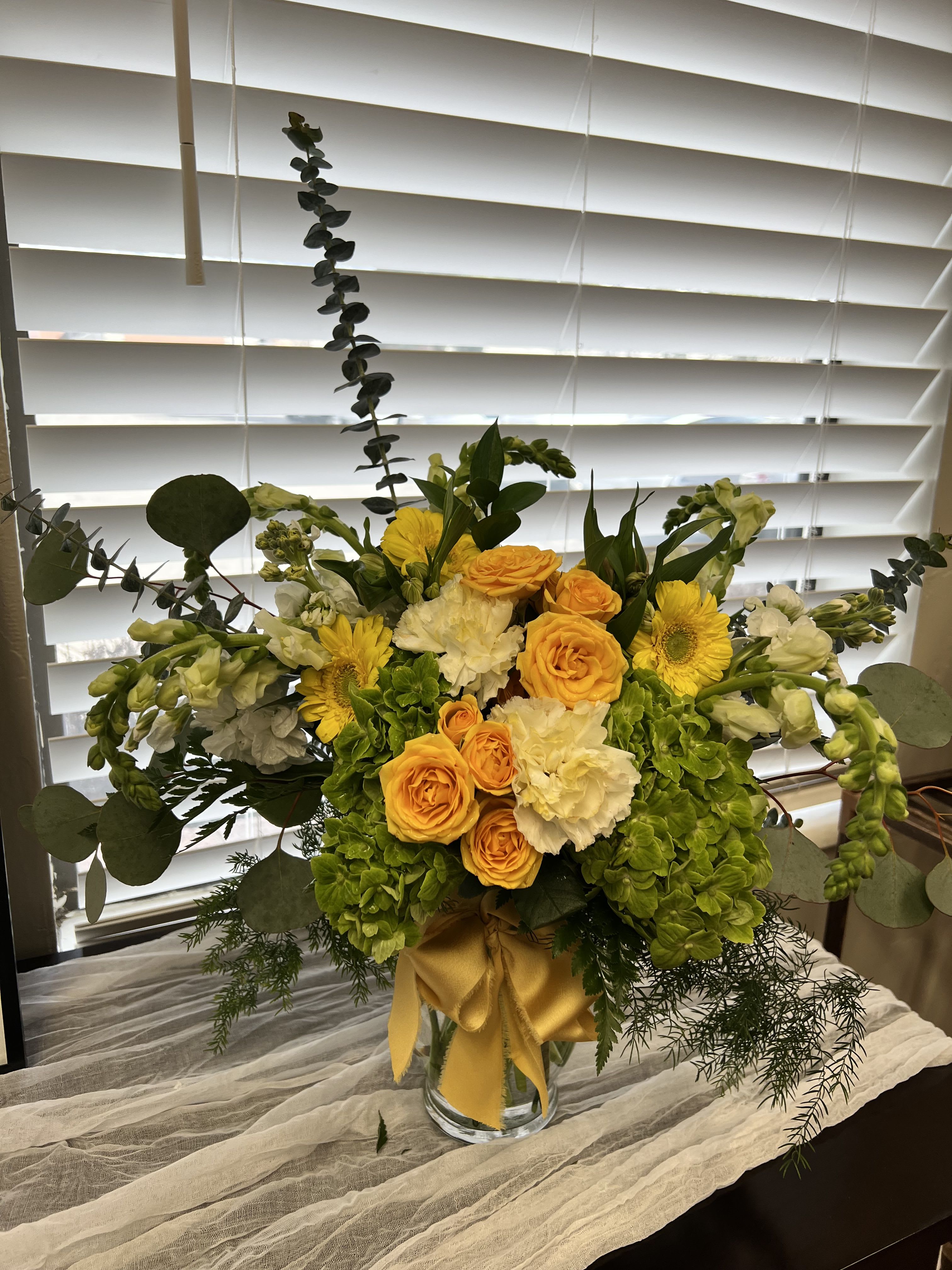 GOLDEN GARDEN RADIANCE  - Bright, joyful, and effortlessly elevated, Golden Garden Radiance is designed to bring warmth and energy into any space. Featuring vibrant golden roses, cheerful gerbera daisies, lush green hydrangea, and soft white stock and carnations, this arrangement is a fresh celebration of color and texture. Layered with flowing eucalyptus and seasonal greenery, the design has a natural, garden-inspired feel while still maintaining a refined, upscale look. The golden satin ribbon detail adds a polished finish, making this piece perfect for birthdays, celebrations, thank-you gifts, or simply brightening someone’s day. Handcrafted with premium blooms and artistic intention, each arrangement is uniquely styled—because sending flowers should feel just as special as receiving them.