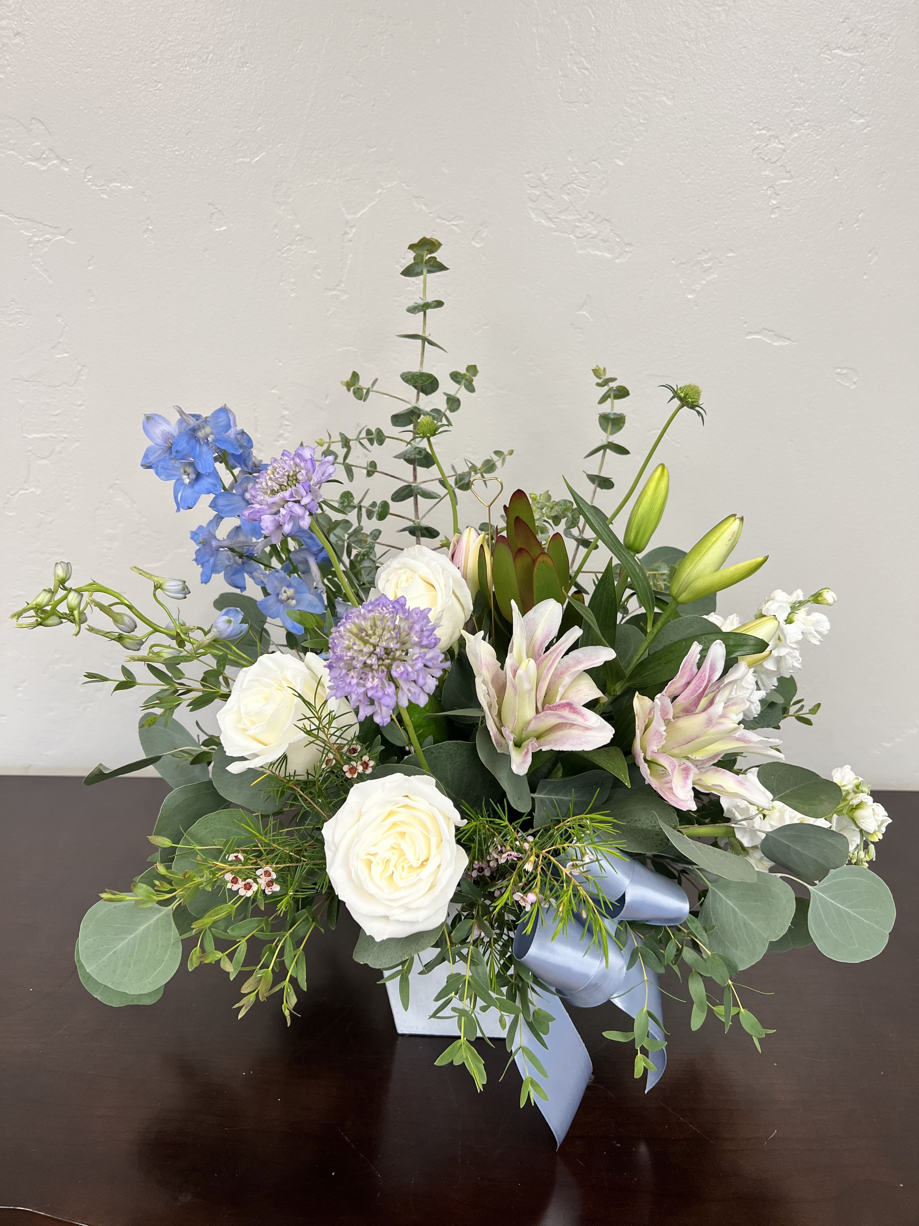 Whispering Garden Elegance - A graceful blend of soft pastels and fresh greenery, Whispering Garden Elegance captures the beauty of a blooming garden in its most serene moment. Featuring premium white roses, fragrant  lilies, delicate lavender scabiosa, and airy blue delphinium, this arrangement is thoughtfully designed with lush eucalyptus and textural accents for a naturally refined look. Presented in a modern vessel with a soft satin ribbon, this floral design offers a perfect balance of elegance and organic charm—ideal for any day! Handcrafted with seasonal premium stems, each arrangement is unique and designed to impress with its fullness, movement, and timeless beauty.