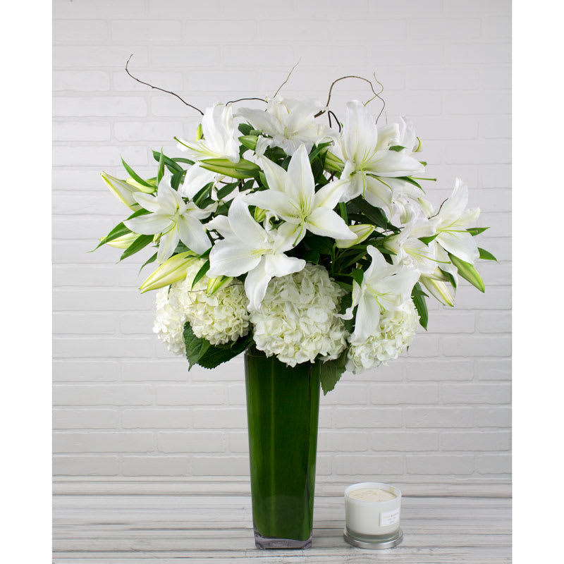 Loving Lilies (DL) - Indulge in the pure and captivating beauty of our large, showy all-white flower arrangement. This stunning ensemble features oriental white lilies and white hydrangeas, accented with curly willow, all artistically arranged in a leaf-lined cylinder vase.  