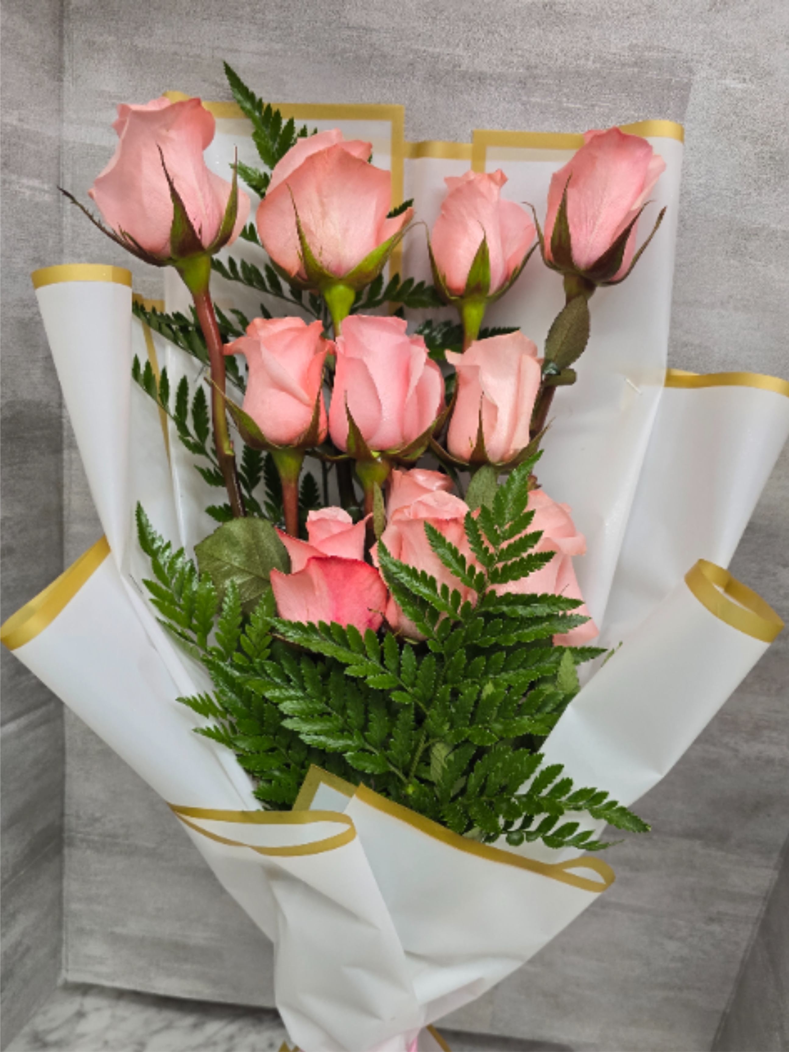 Pink Passion  - Pale pink roses gathered in a loose, hand-tied cluster make this flower arrangement a soft, romantic choice for an anniversary, birthday, or a thoughtful everyday gesture. The "birthday bouquet" or "anniversary roses" feel is immediate - she'll feel adored and noticed the moment the pink petals and feathery greenery are revealed.  The bouquet pairs several pink roses with dense, fern-like foliage for a textured, garden-fresh look. Blooms sit upright in a compact arrangement and are wrapped in white decorative paper with gold edging, then finished with a pink satin ribbon for an elegant touch. White, green and a hint of yellow accents keep the palette light and flattering.  Perfect for celebrating a partner, close friend, or someone you want to make feel truly seen, this arrangement arrives ready to present and makes a graceful impression on the kitchen table or bedside. For best enjoyment, recut stems and place in fresh water upon arrival to extend vase life.