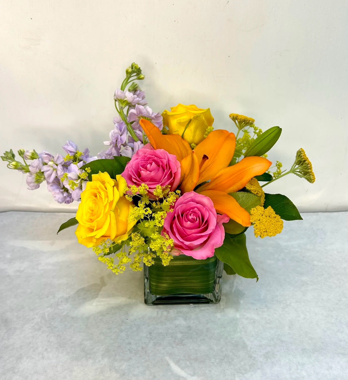 Spring Fling - Vibrant eye catching mix of bright spring flowers in pinks and yellows nestled together in a large clear square vase with a variety of greens and aspidistra leaf accent. HFS 201701-15