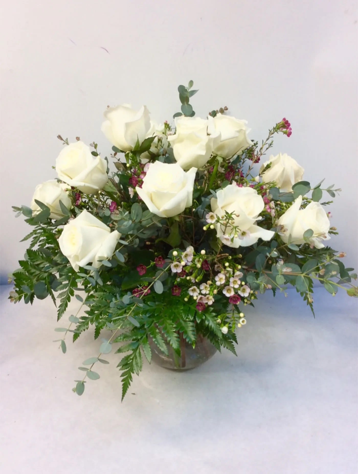 White roses arranged in a glass vase with mixed greenery and small accent flowers.
