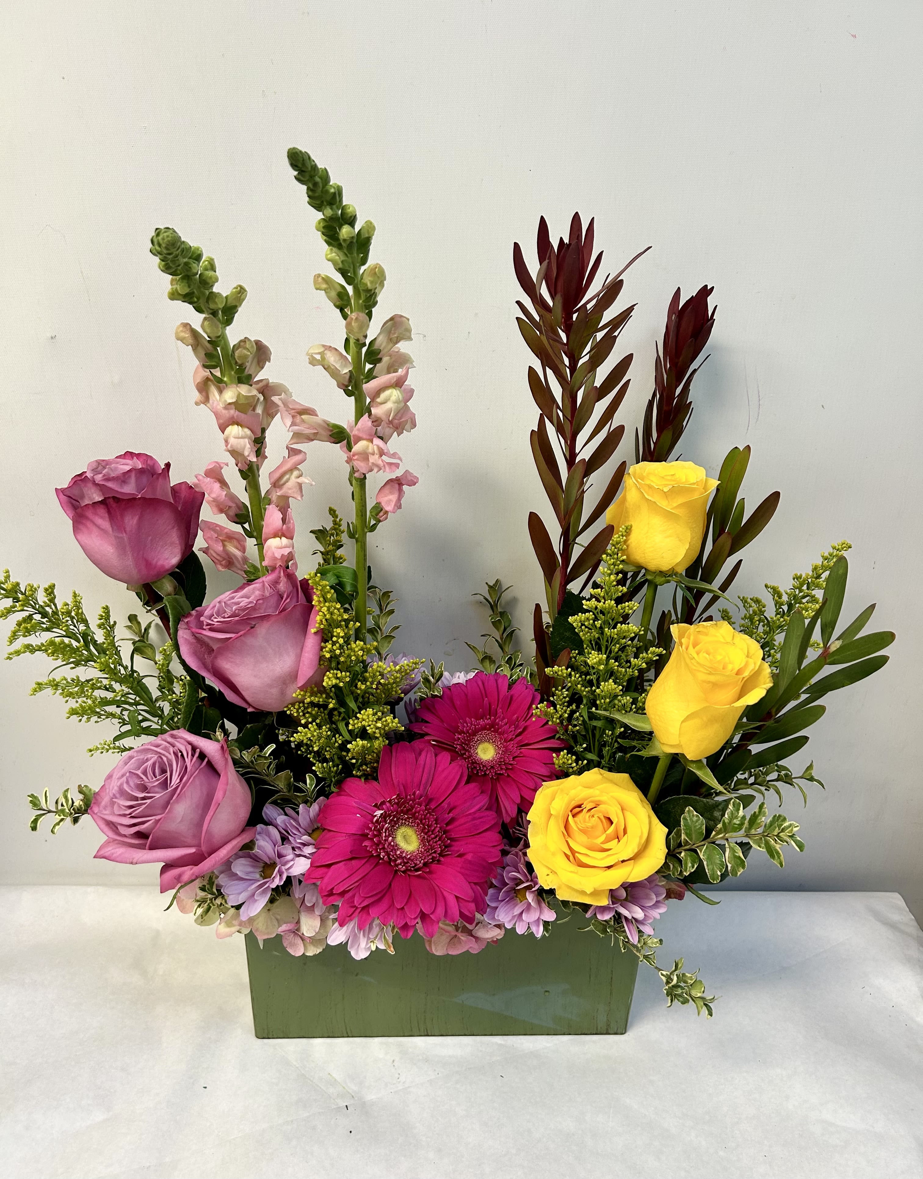 Rectangular arrangement of lavender roses, bright pink gerbera daisies, and yellow roses in a green container