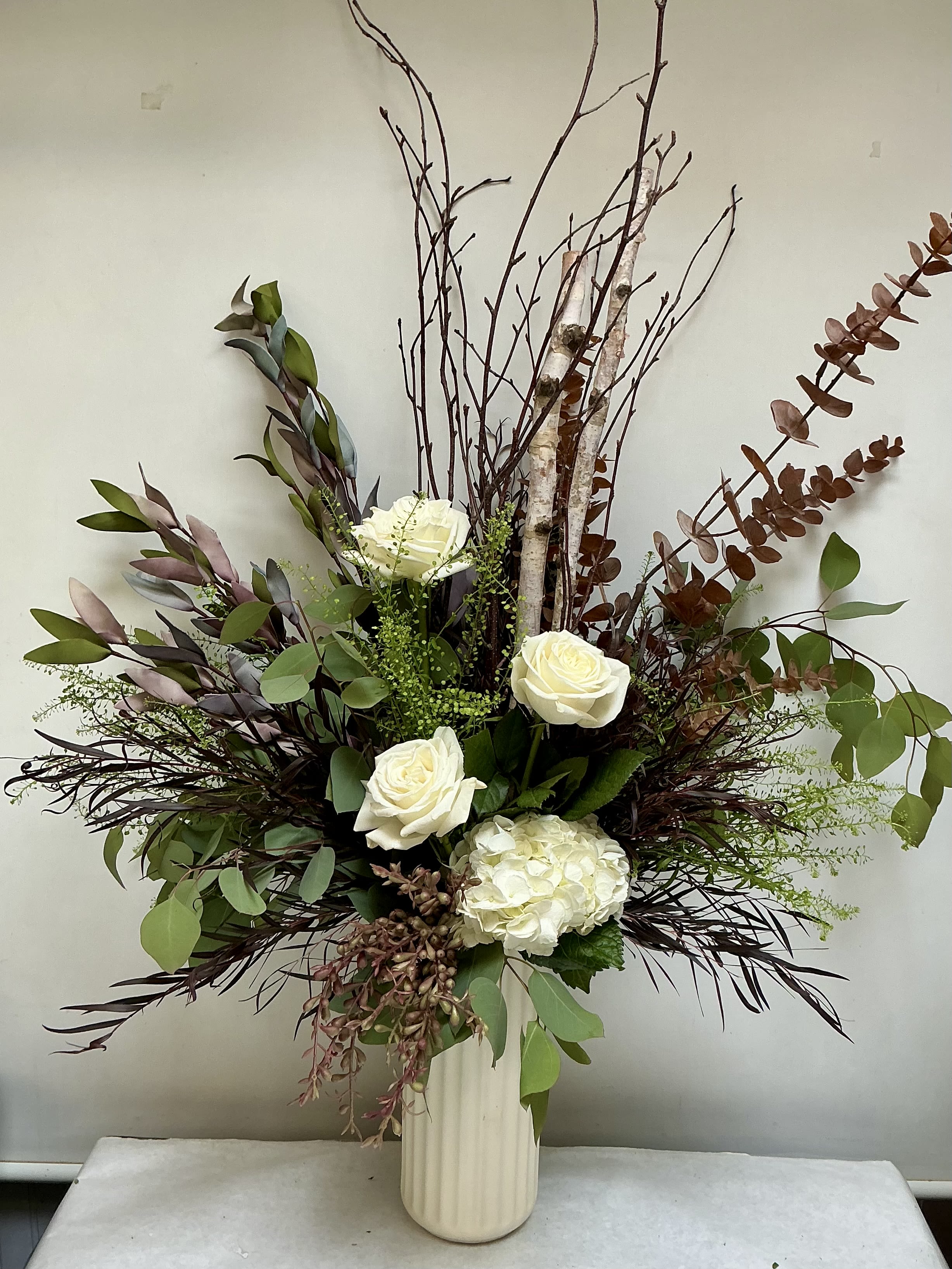 Northwoods Modern - White roses and a single white hydrangea take center stage in this Northwoods Modern flower arrangement, a refined sympathy and funeral bouquet that also suits autumn and Thanksgiving settings. The tall cream ceramic vase and towering birch branches create an airy, asymmetrical silhouette that reads both modern and naturally rustic-perfect for the person who appreciates understated elegance and calm presence.  Deep burgundy foliage and muted green leaves frame the white blooms, while layered greenery and textured birch stems add height, movement, and visual depth. The palette includes accents of red, peach and purple tones for a softly mixed-color effect that keeps the arrangement from feeling stark. The ceramic vase gives a solid, gallery-ready base while the open, expansive design allows the flowers and branches to be admired from every angle.  Intended as a comforting, graceful gift, this piece makes someone feel quietly supported-appropriate for a memorial service or to bring warmth to an autumn table. The arrangement arrives ready to display in its cream ceramic vase; keep in fresh water and out of direct heat to preserve the blooms for as long as possible.