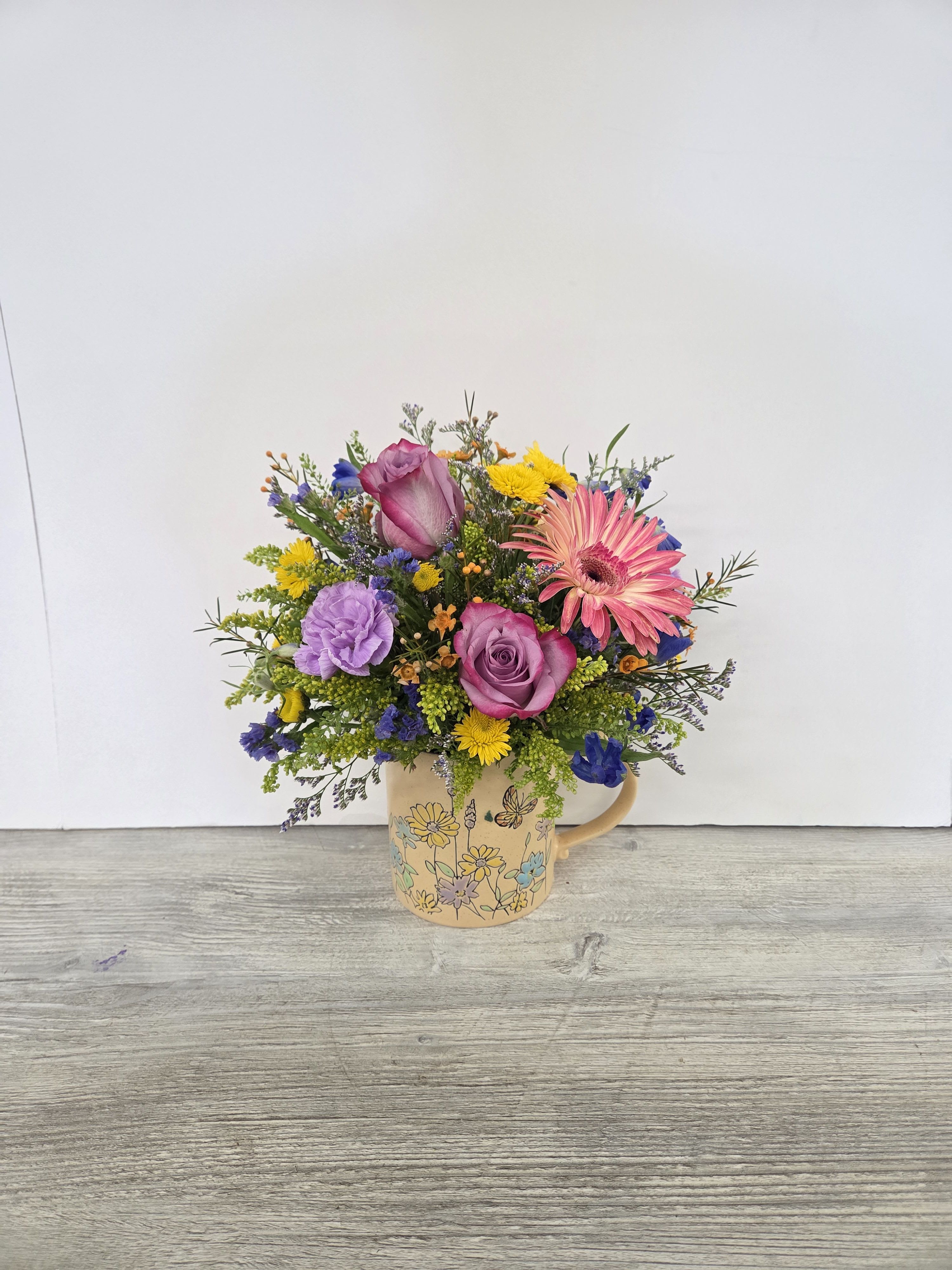 Colorful mixed flower arrangement with pink gerbera, roses, and purple blooms in a floral ceramic mug