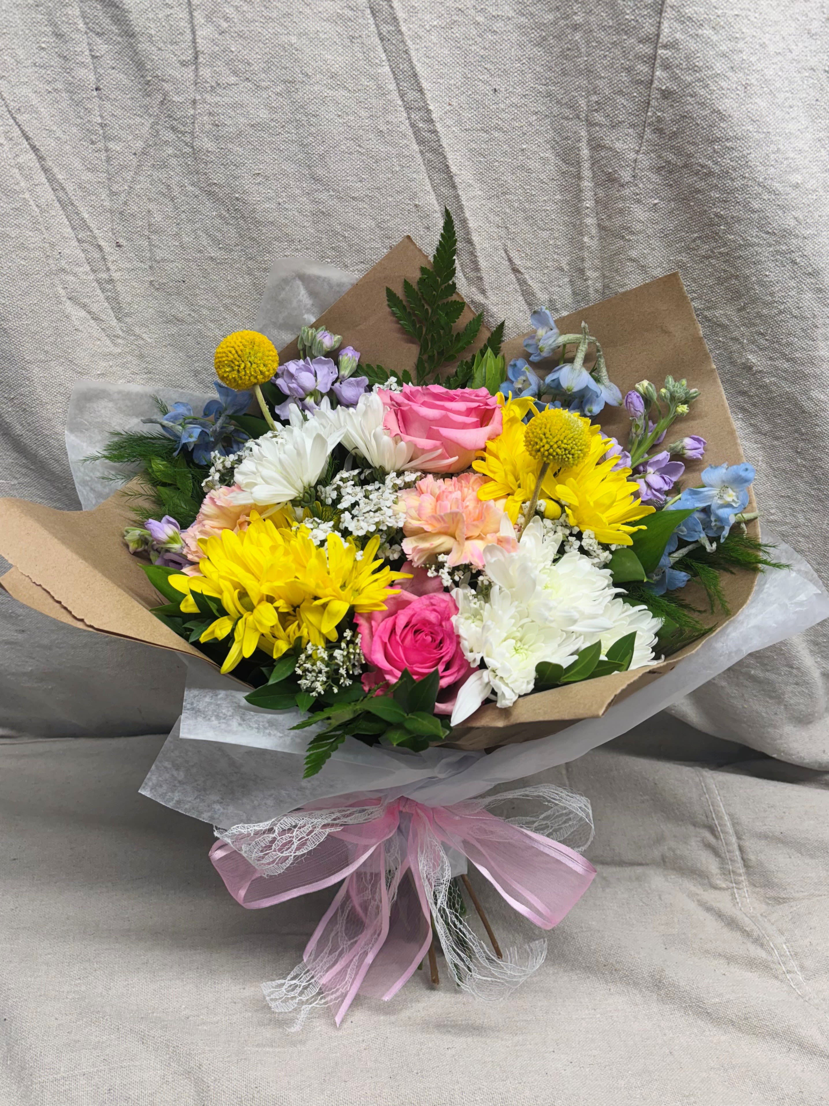Hand-tied bouquet of pink roses, yellow daisies, and white mums wrapped in kraft paper with a pink bow.