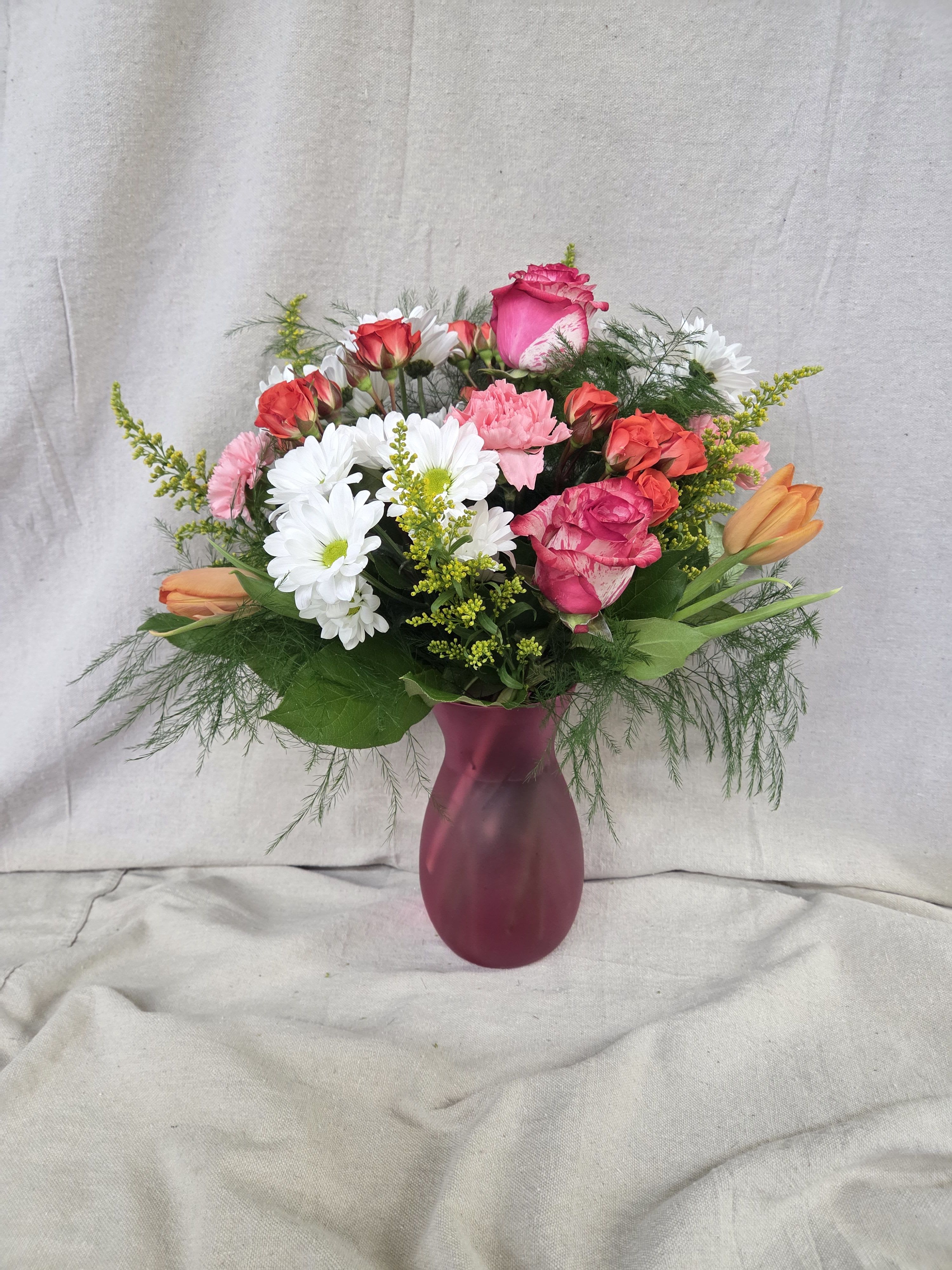 Mixed bouquet of pink roses, white daisies, and orange tulips in a frosted pink vase