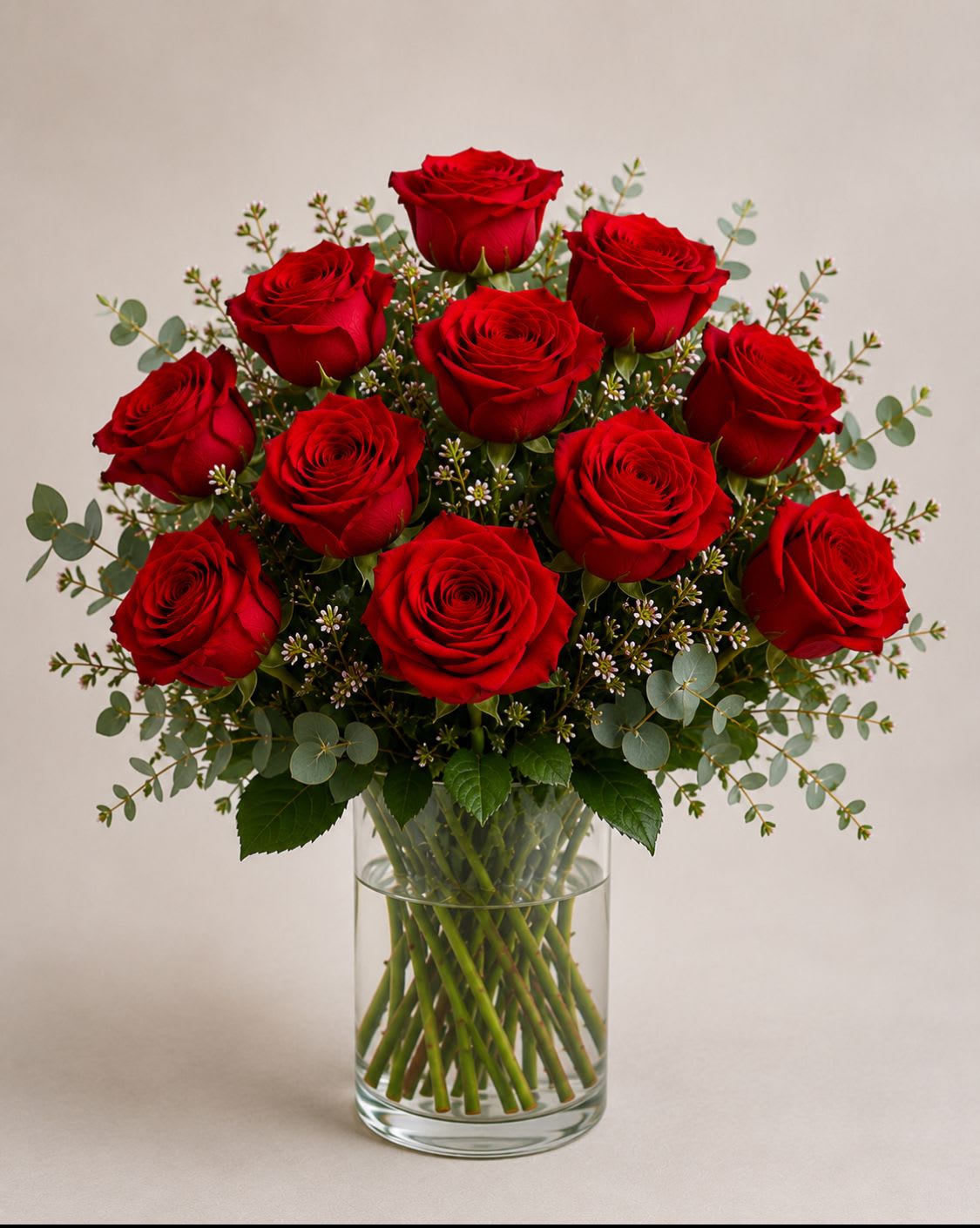 Classic Red  - A dense cluster of red roses gathered in a clear glass vase creates a powerful romantic statement - perfect as a birthday bouquet, anniversary roses, or a Valentine's Day gift for the person you want to make feel truly cherished. The Classic Red arrangement reads formal and elegant, sending a clear message of love and celebration the moment it arrives.  Deep red roses are arranged in a rounded, full shape and softened with airy eucalyptus and small white filler blooms for contrast and texture. Stems are visible through the cylindrical clear glass vase, giving a clean, structured presentation that suits a dining table centerpiece or a bedside display. A timeless choice for someone who appreciates classic beauty, this arrangement arrives ready to display; keep water fresh and trim stems every few days to extend bloom life.