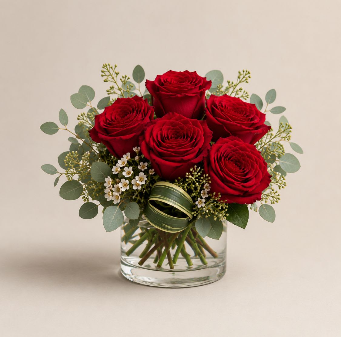 Scarlet - Five deep red roses take center stage in this romantic flower arrangement, perfect for anniversaries, Valentine's Day, or any "thinking of you" moment when you want someone to feel deeply loved. The compact, rounded design reads classic and intimate-an elegant choice for birthdays, Mother's Day, or as a sincere "I'm sorry" gesture-and also appropriate for sympathy or funeral settings when understated beauty is needed.  Arranged in a clear glass vase with a tasteful green ribbon accent, the roses are framed by soft eucalyptus foliage and delicate white filler flowers that add lightness and contrast. The arrangement's size and neat silhouette make it easy to display on a bedside table, office desk, or dining table; water the stems regularly and trim ends every few days to keep the bouquet fresh. This is the kind of bouquet that will make the person you're thinking of feel truly seen and cared for.