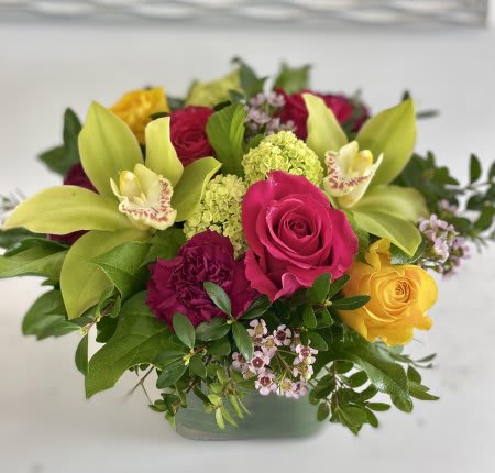 Low arrangement of pink and yellow roses, green orchids, and lime blooms in a glass cube vase