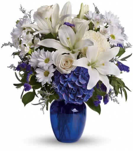White lilies, roses, and daisies with blue hydrangea in a cobalt blue glass vase.