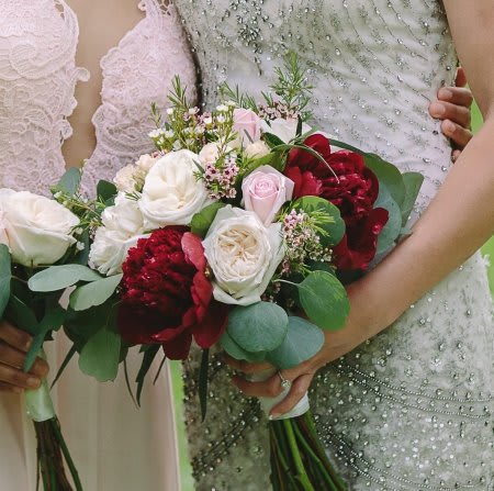 Two women hold bridal bouquets of white, pink, and deep red flowers.