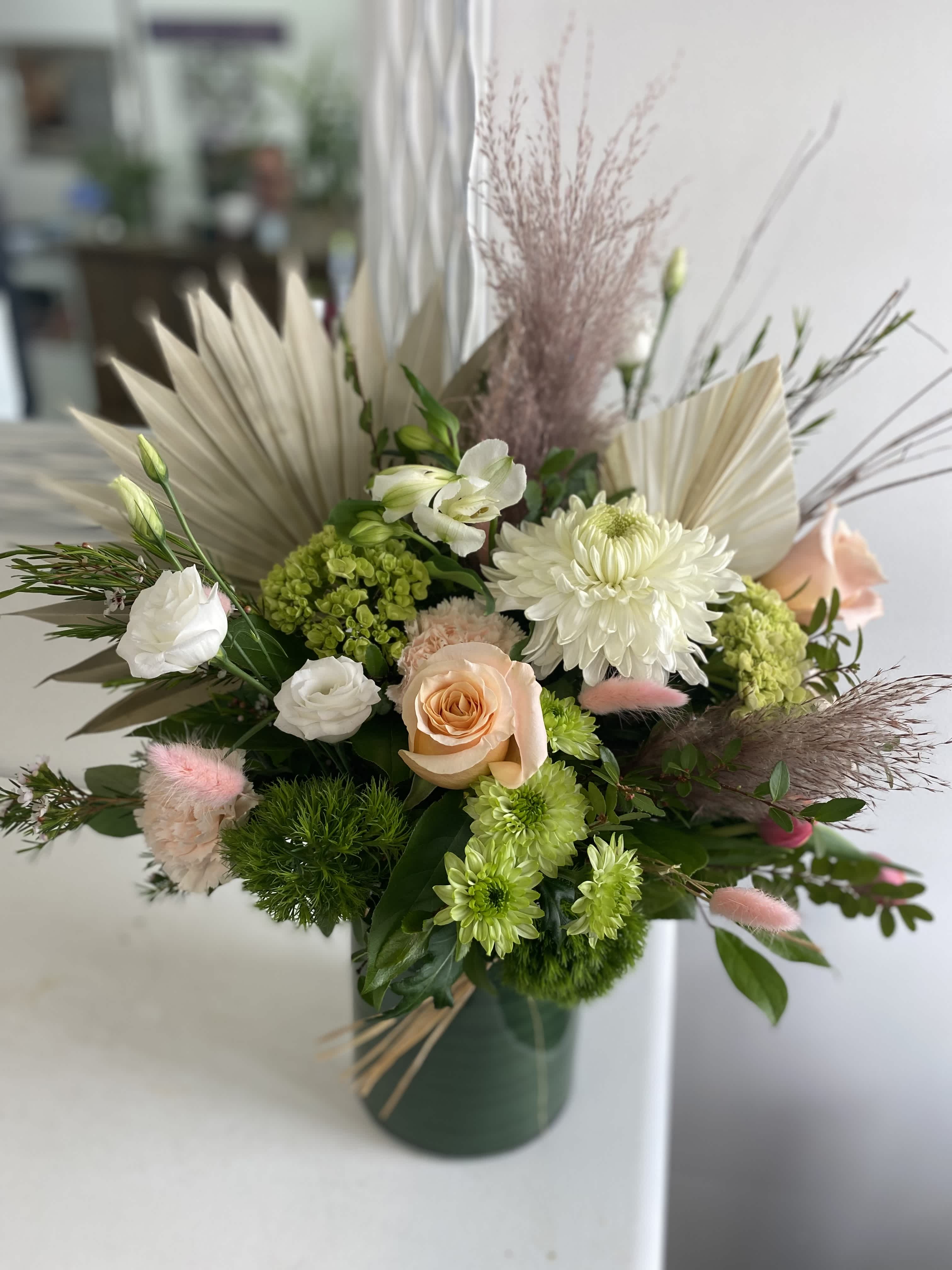 Asymmetrical arrangement of white and peach blooms with green accents in a dark green vase with dried palm fans.