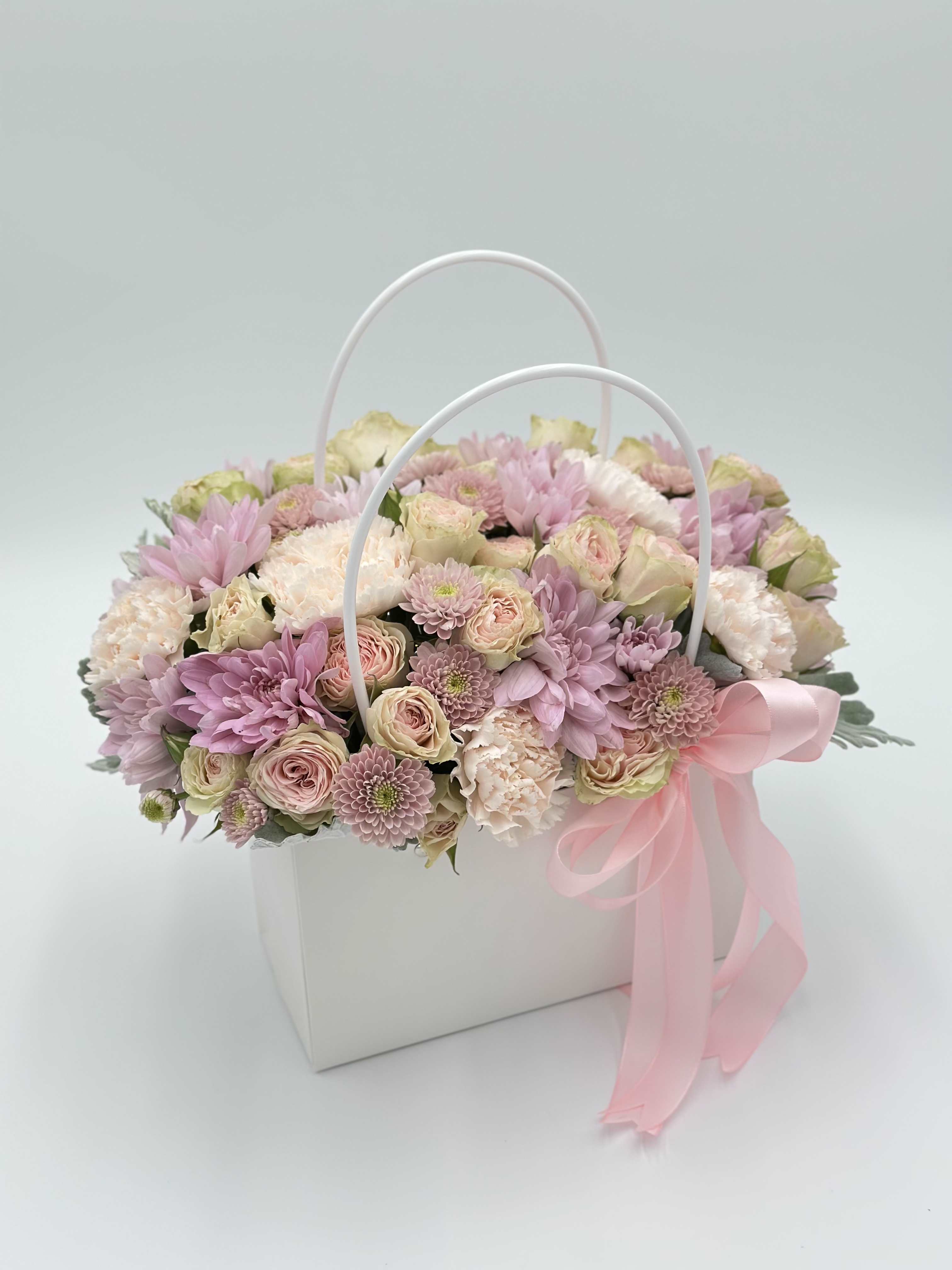 Pink and cream flowers arranged in a white handled box with a pink ribbon