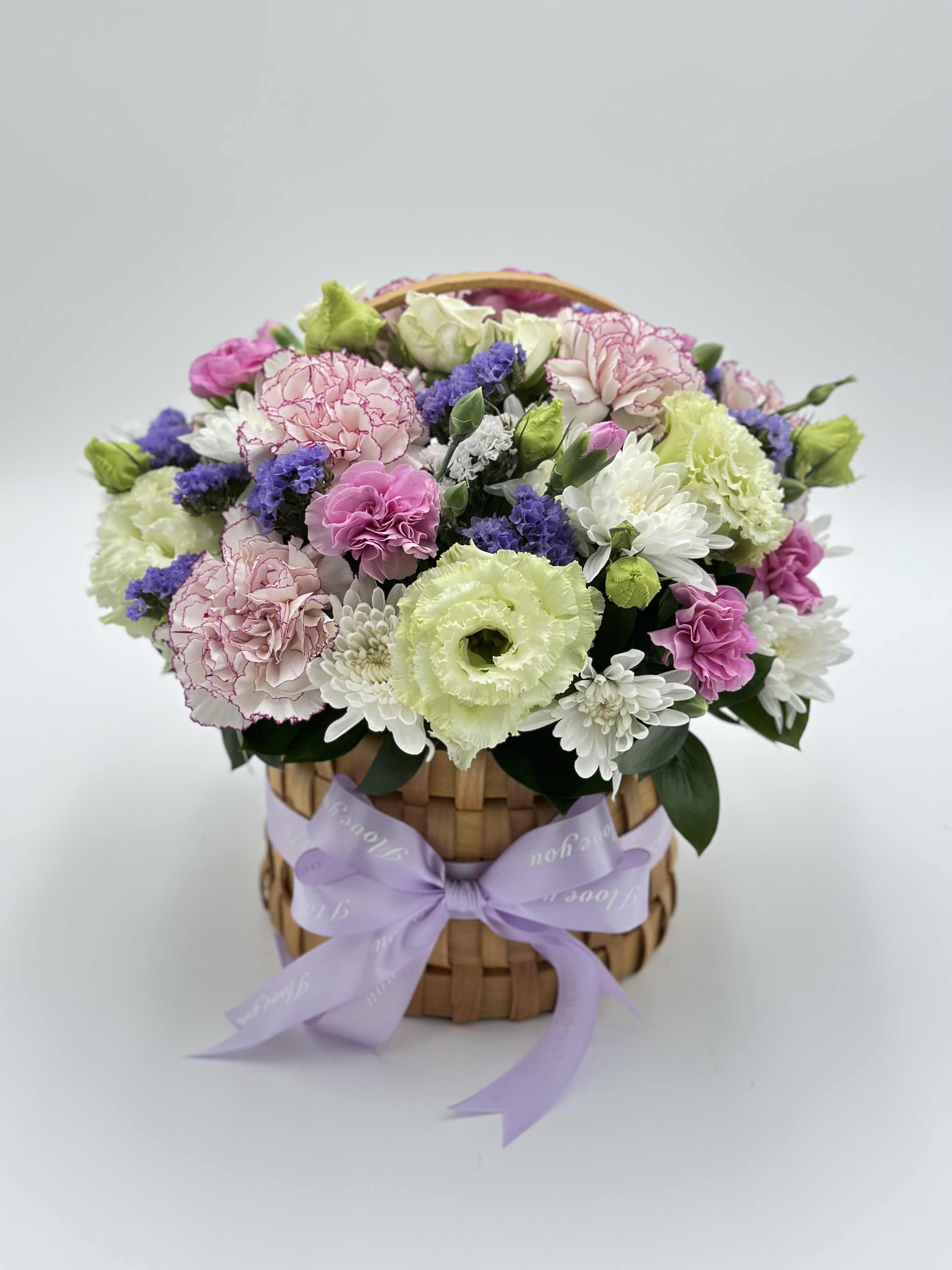 Mixed pink, white, purple, and green flowers in a woven basket with a lavender ribbon