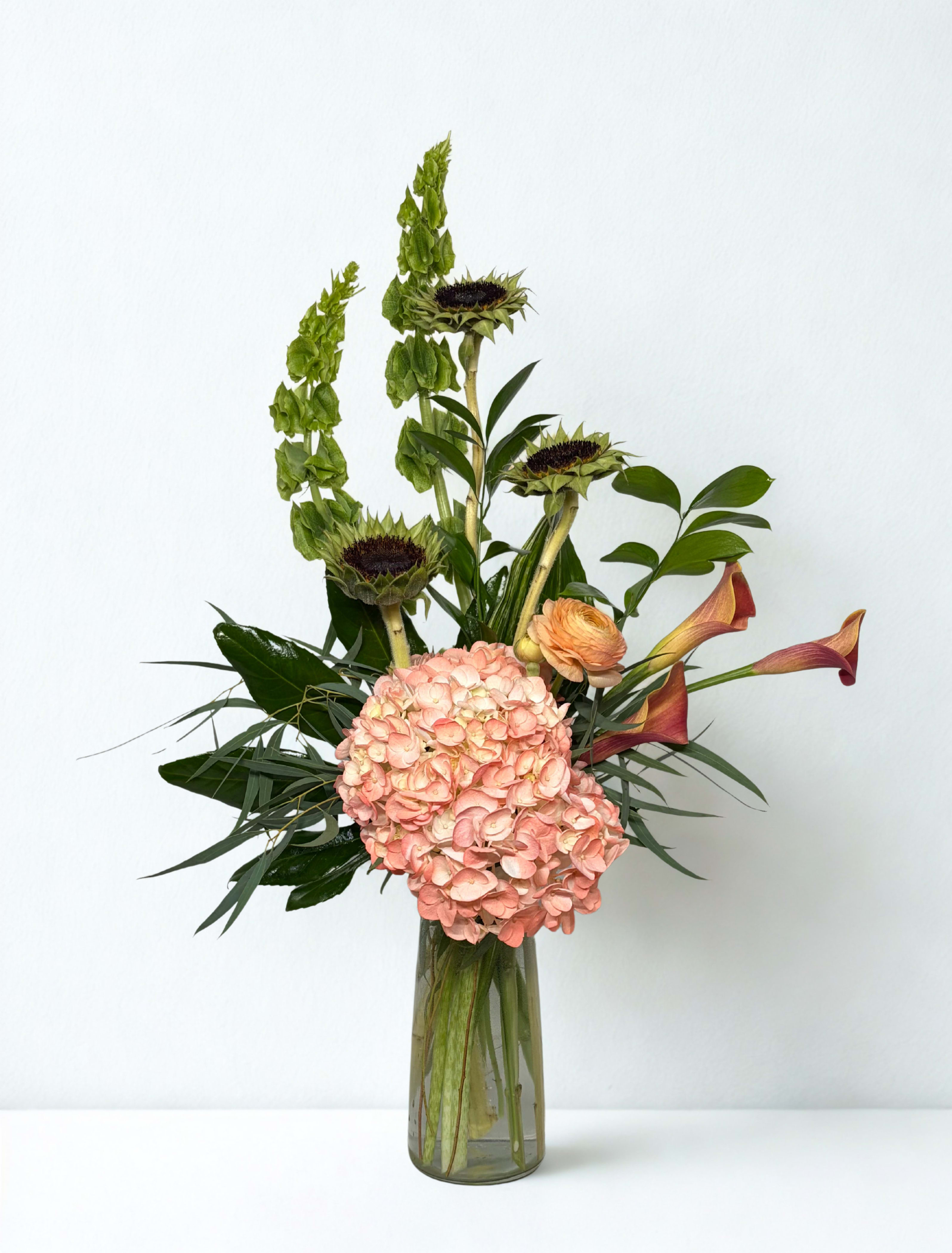 Tall floral arrangement with pink hydrangea, sunflowers, calla lilies, and a glass vase