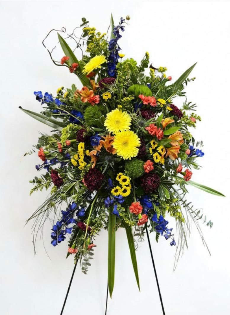Kaleidoscope Garden - A tall, asymmetrical standing spray that greets the room with a burst of mixed-color energy - perfect as a sympathy or funeral tribute or as a bold birthday statement. The arrangement's daisy-like gerbera focal flowers and vivid cobalt delphinium create a bright, uplifting presence that honors the moment while celebrating a life or milestone.  Closely clustered carnations, chrysanthemums, alstroemeria and daisy accents in yellow, orange, green and burgundy give layered texture and lively contrast, while long arching foliage adds graceful height on the easel. Designed for visibility at a service or front-and-center at a party, it arrives ready to display on its standing easel; light water tubes keep blooms fresh until placed.   As similar as possible. Designer may make appropriate substitution based on flower availability. 