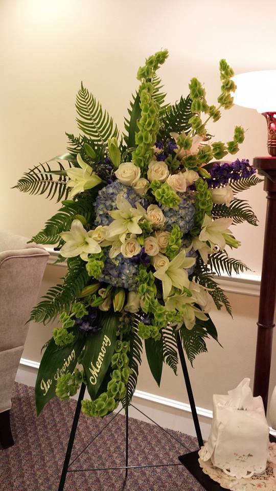 Tall standing spray of white lilies, roses, blue hydrangeas, and green bells of Ireland on an easel