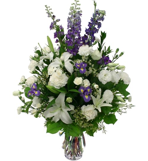 Tall arrangement of white lilies and carnations with purple delphinium and iris in a clear glass vase