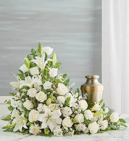 Asymetrical Cremation Arrangement- All White - Our all-white arrangement sends a heartfelt message of hope to those coping with loss. Handcrafted in an elegant asymmetrical design, it’s a beautiful tribute to honor the life of someone very special.One-sided arrangement of white roses, Asiatic lilies, snapdragon, carnations, stock and monte casino; accented with assorted greenery