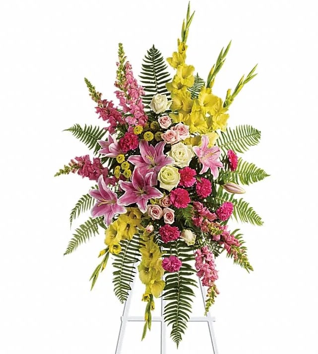 Tall standing spray with pink lilies, roses, carnations, yellow blooms, and ferns on a white easel.