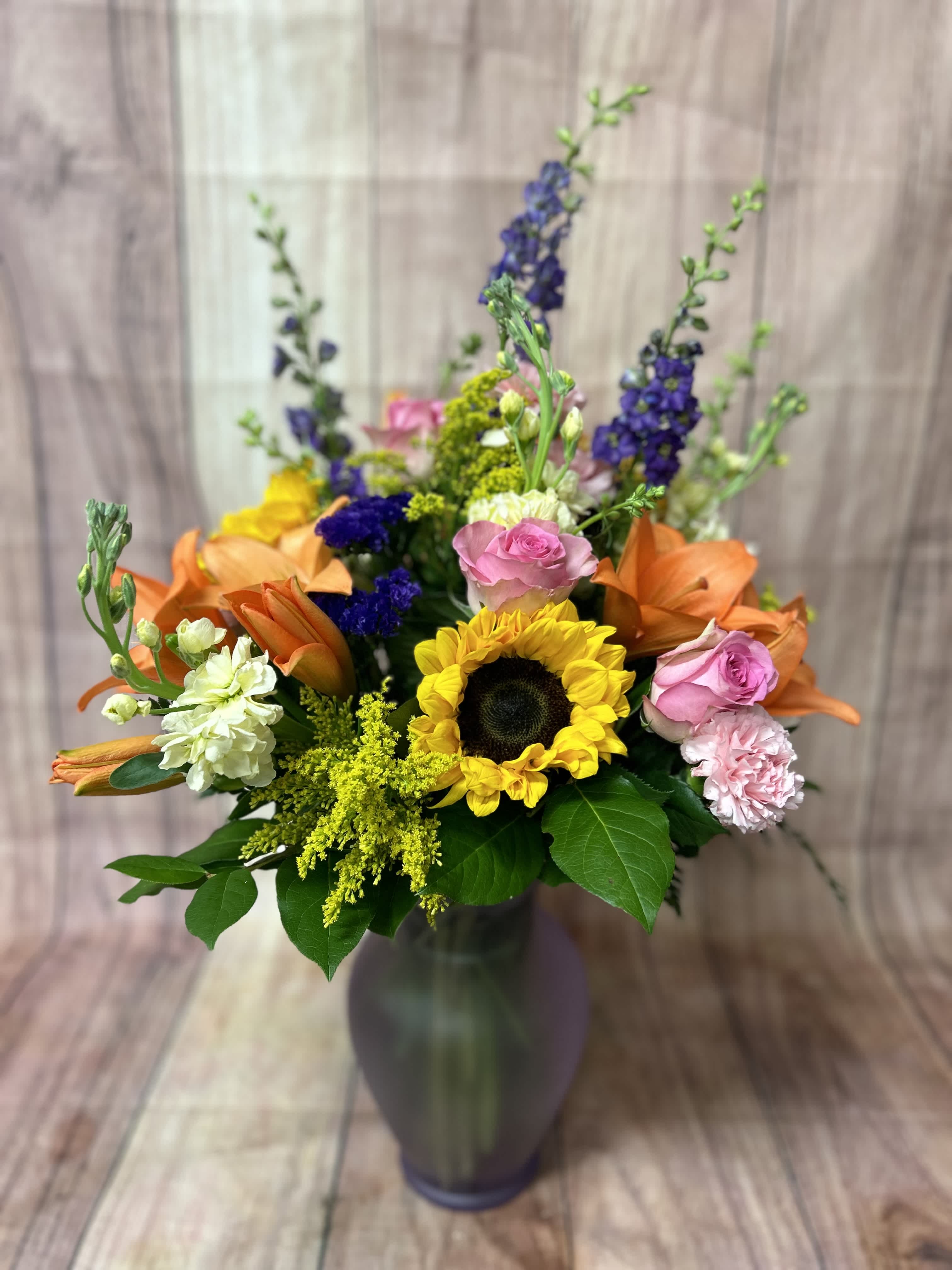 Mixed bouquet in a tall vase with sunflowers, roses, lilies, and purple spikes