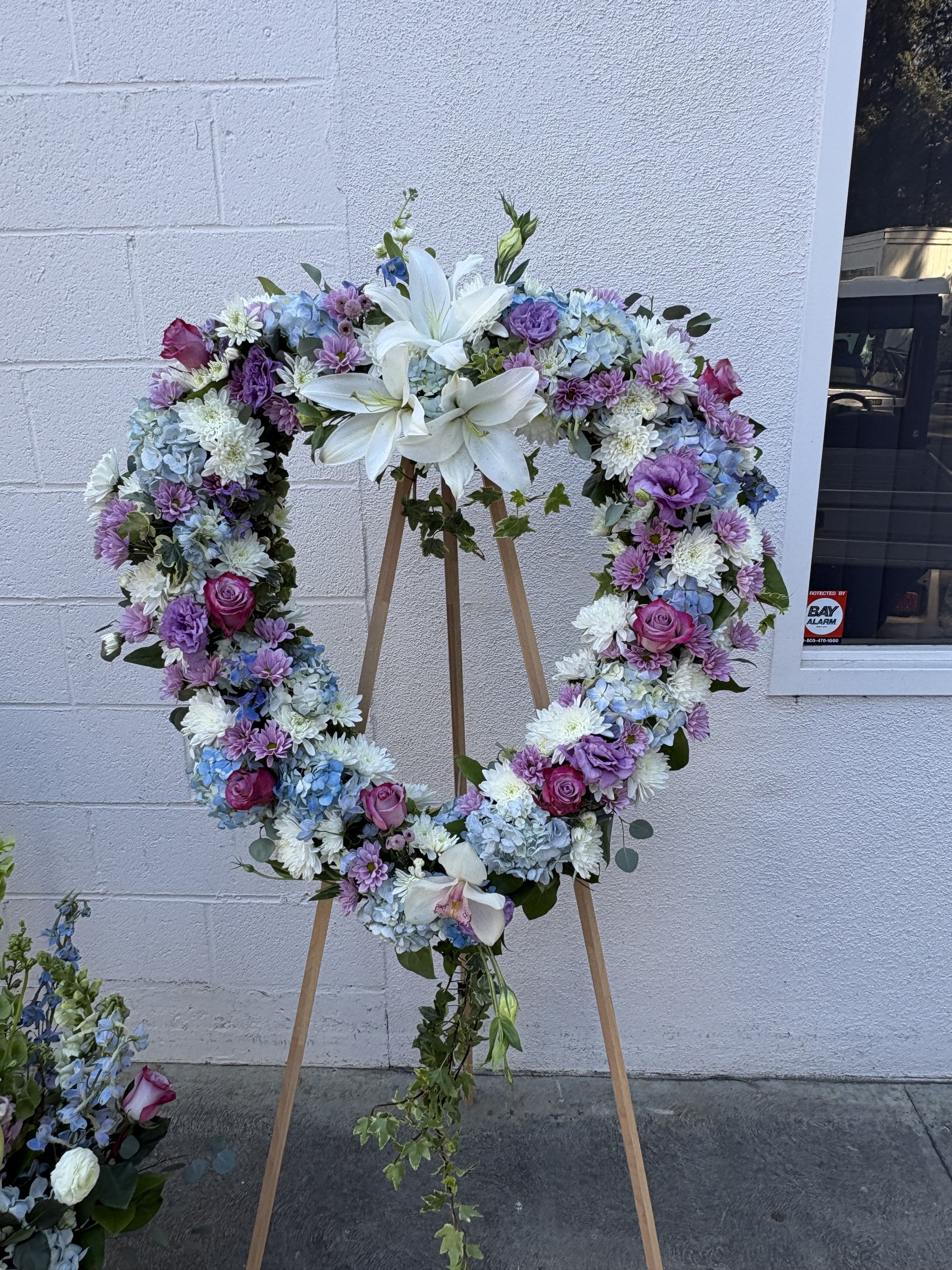 Blue and Lavender Heart-Shaped Sympathy Wreath Easle - Our Blue and Lavender Heart-Shaped Sympathy Wreath Easle includes white casablanca lilies, lavender roses, blue hydrangaes, white daisies, white chrysanhemums, lavender lisianthus, White Cymbidium Orchid with ivy and lisianthus draping down the middle.